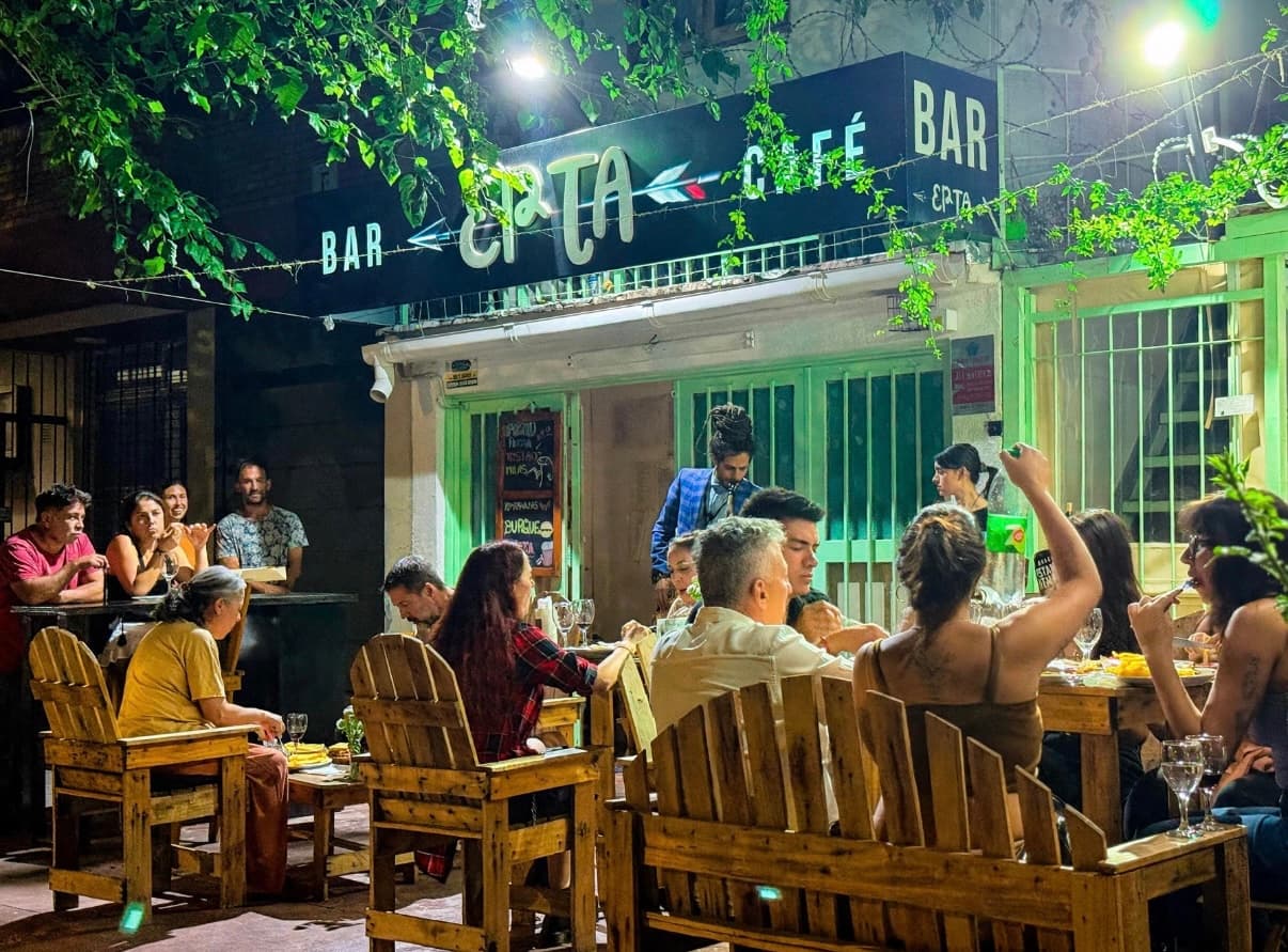 EPTA de noche: el cartel «Bar EPTA Café» iluminado y la vereda llena de gente comiendo en mesas y sillones de madera